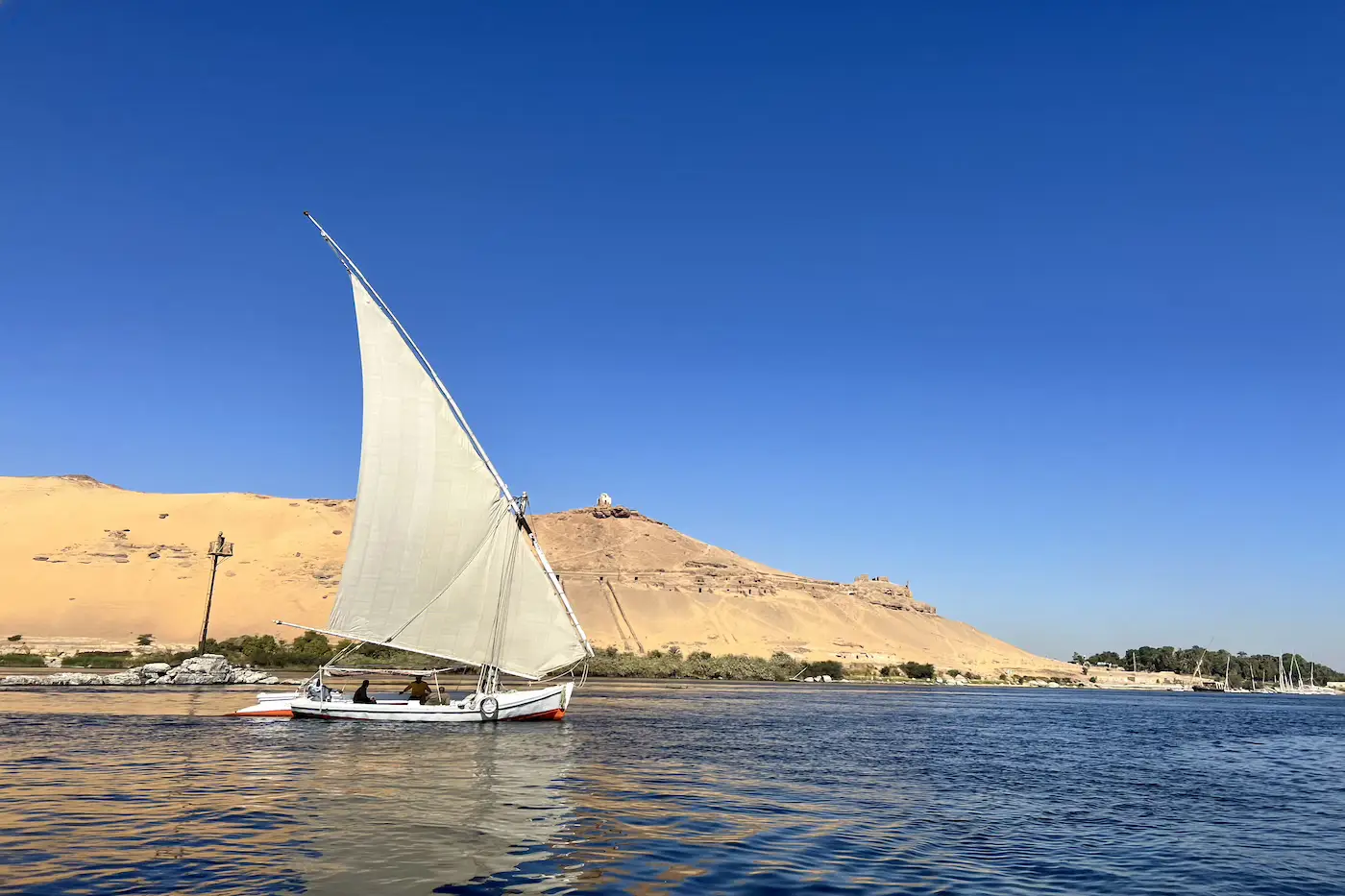 Traditional wooden dahabiya under full sail on the Nile at golden hour with palm-lined banks.